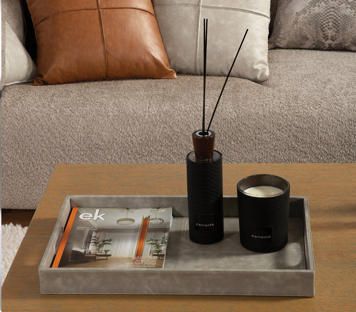Γκρι δερμάτινος δίσκος σε τραπεζάκι σαλονιού, διακοσμημένος με περιοδικό, μαύρο αρωματικό χώρου και κερί. Στο φόντο καναπές με μαξιλάρια σε καφέ, μπεζ και γκρι αποχρώσεις.