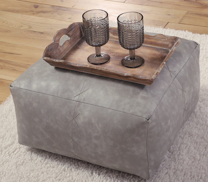 Γκρι πουφ σε μοντέρνο design με διακοσμητικές ραφές, τοποθετημένο σε σαλόνι με ξύλινο δίσκο και ποτήρια.