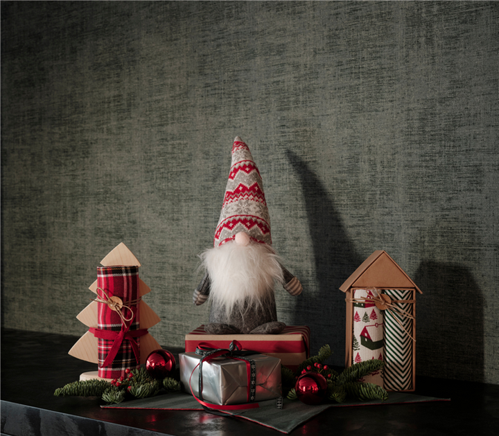 Χριστουγεννιάτικο στοπ πόρτας σε σχήμα νάνου με γκρι σώμα, πλεκτό σκούφο σε κόκκινο και γκρι μοτίβο και μακριά λευκή γενειάδα, τοποθετημένο δίπλα σε γιορτινά δώρα και διακοσμητικά.