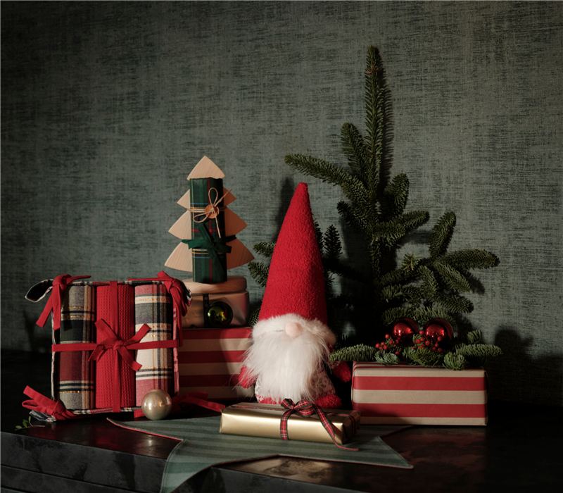 Χριστουγεννιάτικο στοπ πόρτας σε σχήμα νάνου με κόκκινο σκούφο και πλεκτές λεπτομέρειες, διακοσμημένο με δώρα, κουκουνάρια και κλαδιά ελάτου σε γιορτινό σκηνικό.