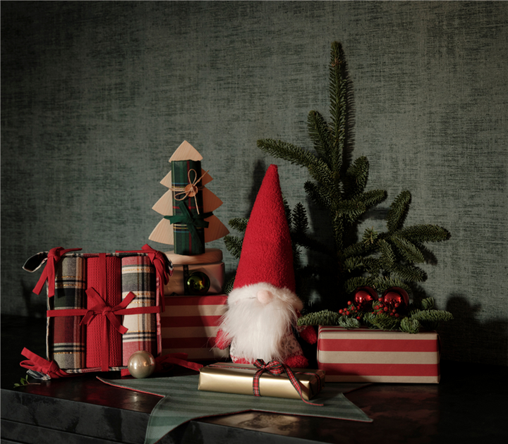 Χριστουγεννιάτικο στοπ πόρτας σε σχήμα νάνου με κόκκινο σκούφο και πλεκτές λεπτομέρειες, διακοσμημένο με δώρα, κουκουνάρια και κλαδιά ελάτου σε γιορτινό σκηνικό.