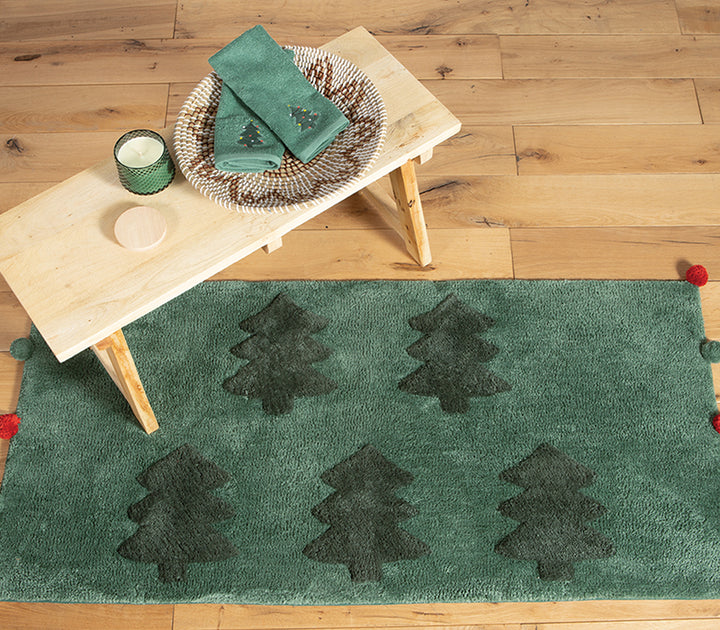 Χαλάκι μπάνιου σε πράσινη απόχρωση με ανάγλυφα δέντρα και κόκκινες-πράσινες φουντίτσες, συνδυασμένο με πετσέτες και κερί για γιορτινή διακόσμηση μπάνιου.