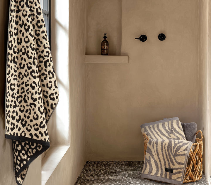 Μπεζ πετσέτα μπάνιου με leopard animal print κρεμασμένη σε μοντέρνο μπάνιο, με καλάθι που περιέχει πετσέτες zebra μοτίβου.