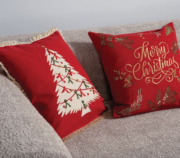 Δύο κόκκινα χριστουγεννιάτικα μαξιλάρια σε καναπέ, το ένα με κέντημα “Merry Christmas” και το άλλο με λευκό δέντρο διακοσμημένο με κορδέλες, σε ζεστό σαλόνι με διακόσμηση εορτών.