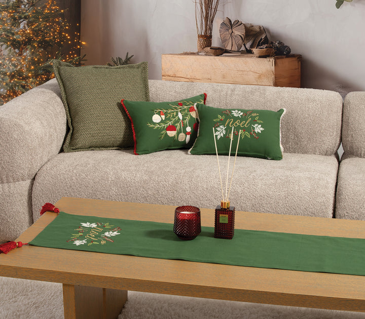 Χριστουγεννιάτικη διακόσμηση σαλονιού με πράσινα μαξιλάρια, τραβέρσα στο ίδιο μοτίβο “Noel”, κόκκινο κερί και αρωματικό χώρου σε ξύλινο τραπεζάκι.