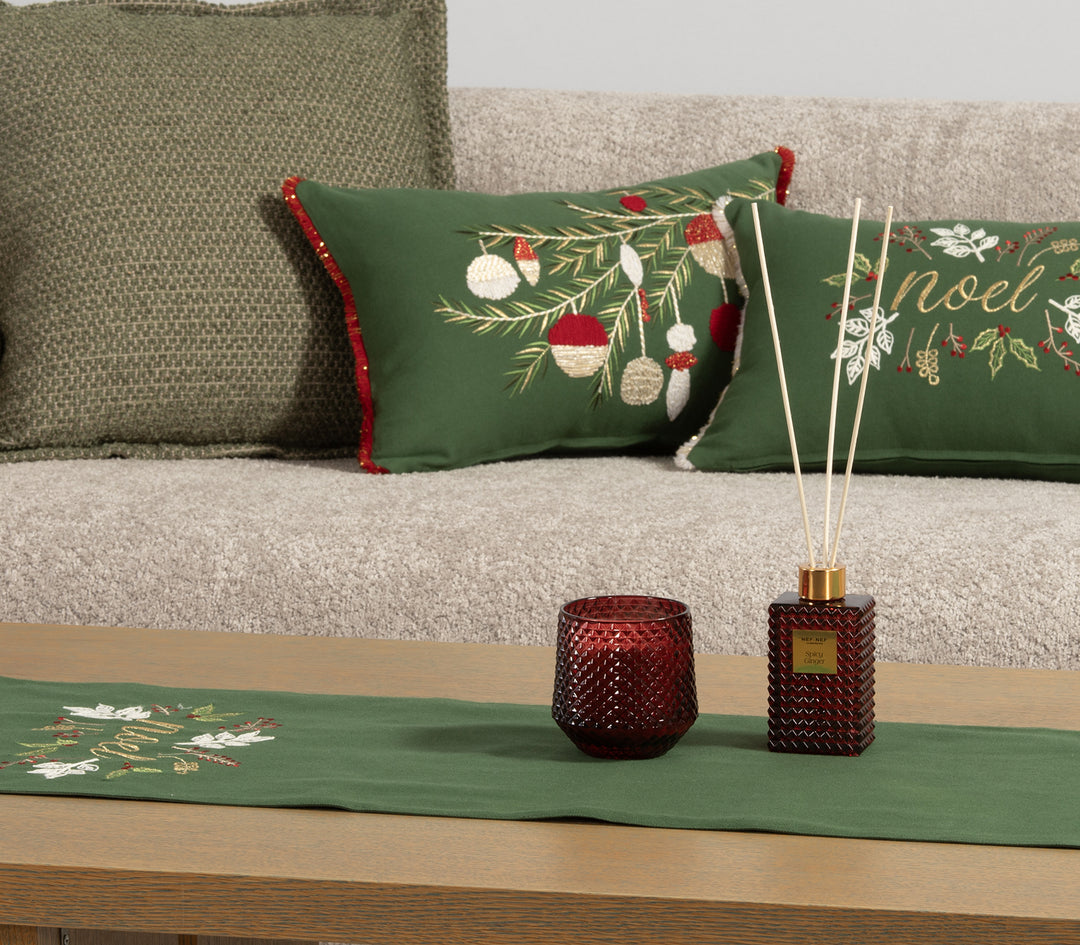 Κοντινό πλάνο από σαλόνι με πράσινα μαξιλάρια χριστουγεννιάτικου θέματος και τραβέρσα με το ίδιο μοτίβο “Noel”, δίπλα σε κόκκινο κερί και αρωματικό χώρου.
