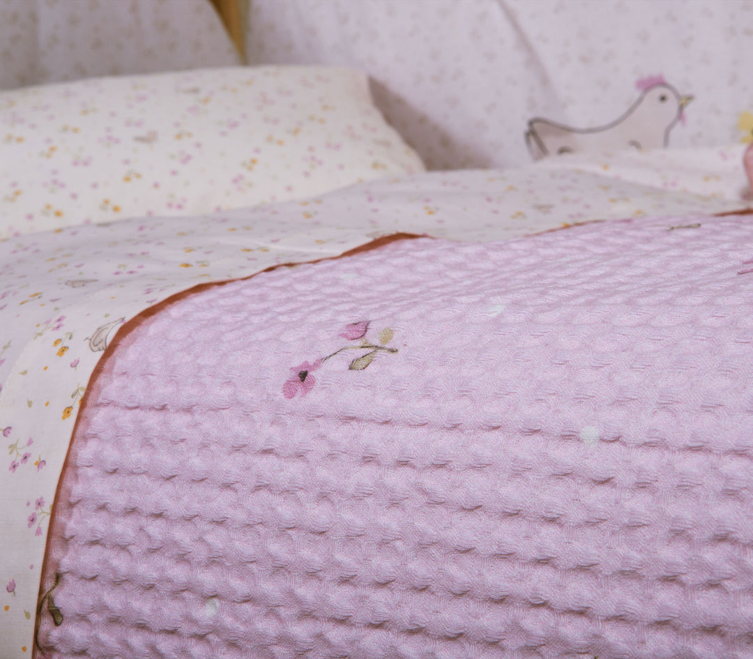 Κοντινή λήψη βρεφικής κουβέρτας πικέ λιλά με μικρό κεντημένο λουλουδάκι τοποθετημένη πάνω σε βρεφικό κρεβάτι.