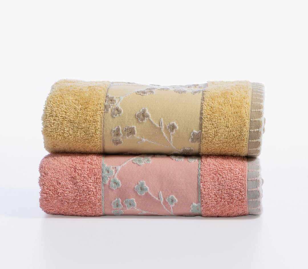 Δύο βαμβακερές πετσέτες Floriva σε κίτρινη και κοραλί απόχρωση διπλωμένες με διακοσμητική μπορντούρα λουλουδιών σε λευκό φόντο.