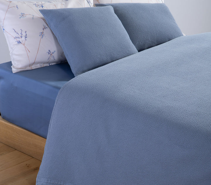 Μπλε διακοσμητικές μαξιλαροθήκες Nef-Nef Homeware πάνω σε κρεβάτι με σεντόνια με λεπτό φυτικό μοτίβο.