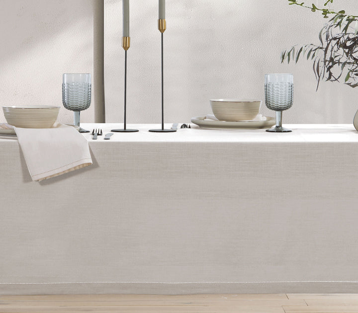 Εκρού βαμβακερό τραπεζομάντηλο στρωμένο σε τραπέζι με σερβίτσια, ποτήρια και διακοσμητικά κεριά σε φωτεινό χώρο.