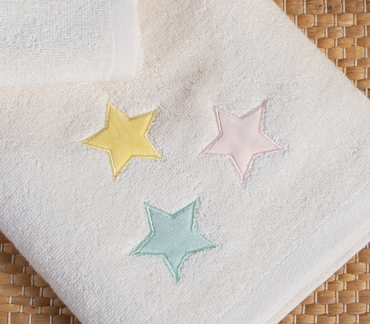 Κοντινή λήψη βρεφικής πετσέτας εκρού με τρία κεντημένα αστέρια σε παστέλ αποχρώσεις πάνω σε ψάθινο φόντο.