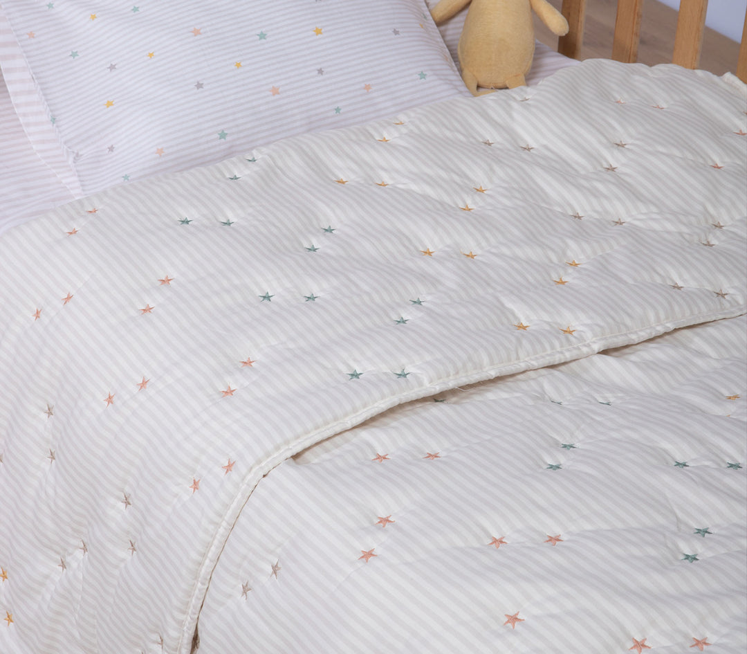 Βρεφικό κουβερλί σε εκρού απόχρωση με μικρά πολύχρωμα αστέρια τοποθετημένο σε βρεφικό κρεβατάκι με μαξιλάρι και λούτρινο παιχνίδι.