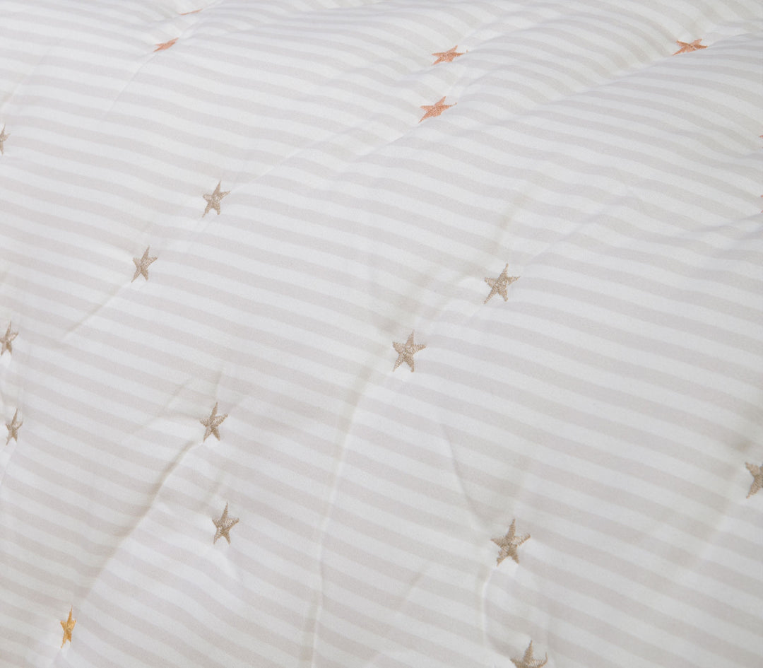 Κοντινό πλάνο βρεφικού κουβερλί με μικρά κεντημένα αστέρια σε απαλές αποχρώσεις πάνω σε εκρού ριγέ ύφασμα.