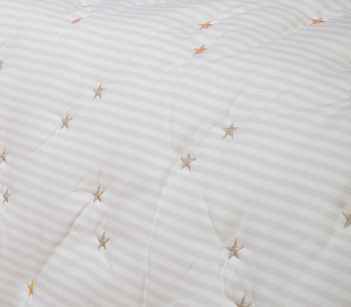 Κοντινό πλάνο βρεφικού κουβερλί με μικρά κεντημένα αστέρια σε απαλές αποχρώσεις πάνω σε εκρού ριγέ ύφασμα.