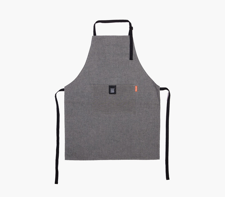 Γκρι ποδιά κουζίνας με μαύρα λουράκια, μπροστινή τσέπη και minimal σχεδιασμό σε ουδέτερο φόντο.