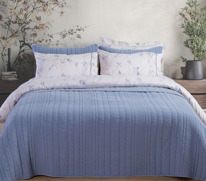 Μπλε καπιτονέ κουβερλί Nef-Nef Homeware σε κρεβάτι με διακοσμητικά μαξιλάρια και φυσικό ξύλινο έπιπλο στο φόντο.