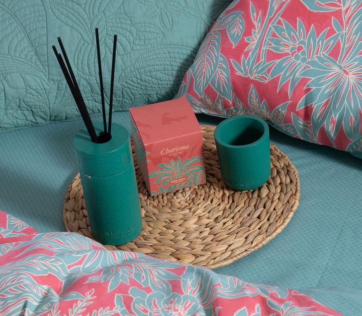 Αρωματικό χώρου και κερί Charisma τοποθετημένα σε ψάθινο δίσκο επάνω σε κρεβάτι με διακοσμητικά μαξιλάρια.