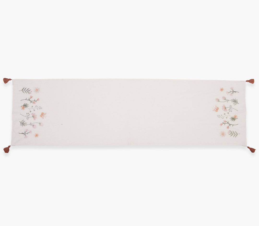 Τραβέρσα σε εκρού-ροζ απόχρωση με διακριτικά λουλουδάκια και φουντάκια στις άκρες σε λευκό φόντο.