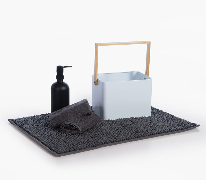Λευκό μεταλλικό καλάθι μπάνιου με ξύλινη λαβή, πετσέτα και δοχείο σαπουνιού τοποθετημένα πάνω σε γκρι χαλάκι μπάνιου.