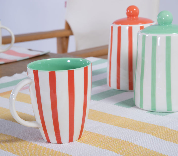Κεραμική κούπα με κόκκινες ρίγες σε τραπέζι με δοχεία και ριγέ σουπλά.
