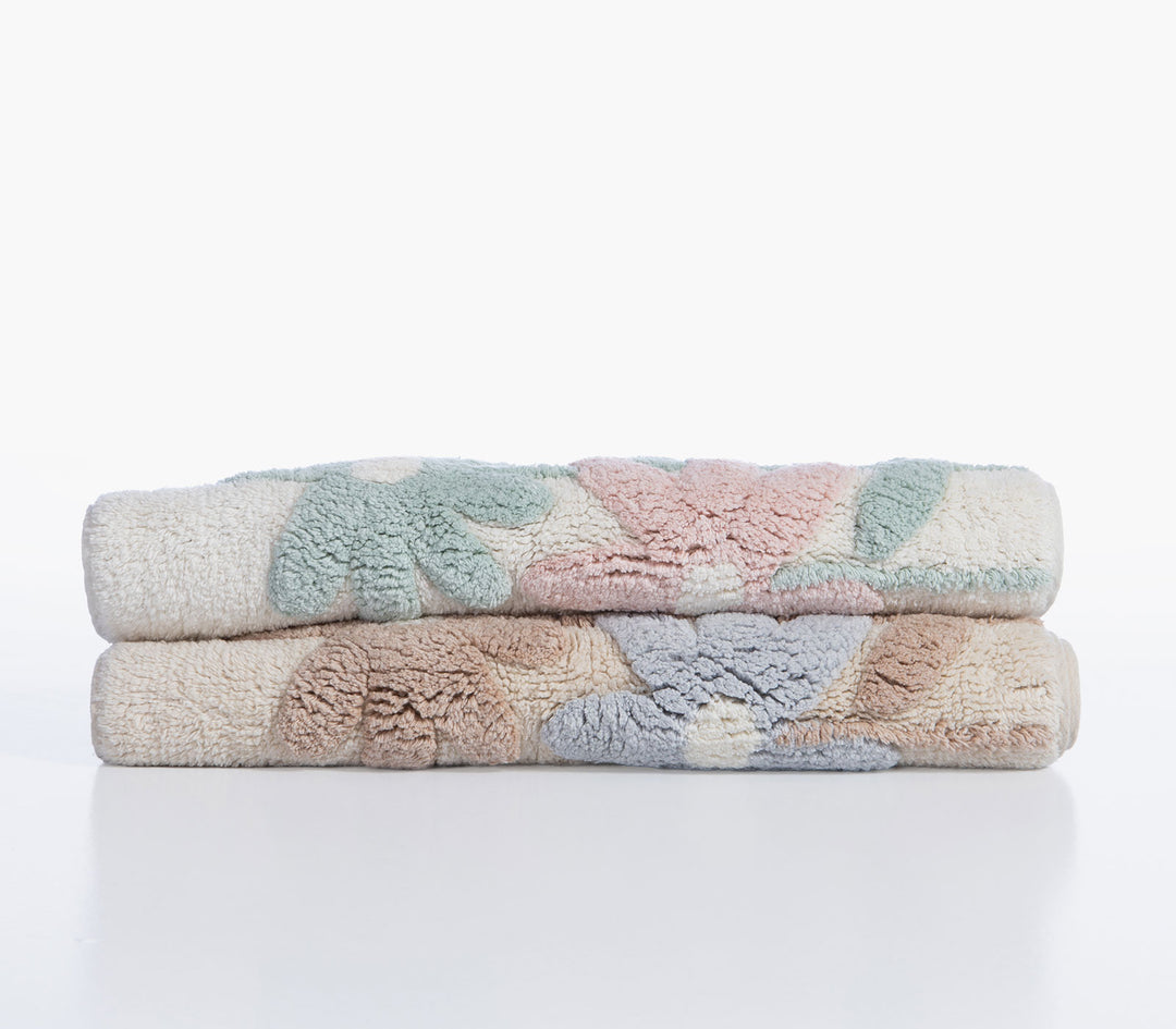 Δύο διπλωμένα βαμβακερά ταπέτα μπάνιου Dreamel με ανάγλυφα λουλούδια σε παστέλ αποχρώσεις πάνω σε λευκό φόντο.
