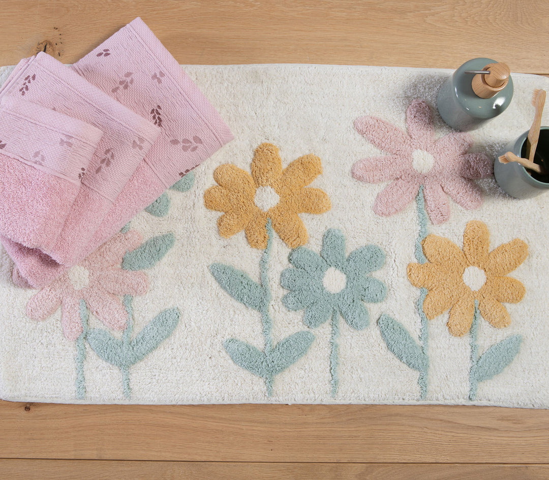 Εκρού ταπέτο μπάνιου με ανάγλυφα παστέλ λουλούδια σε ξύλινο δάπεδο, μαζί με ροζ πετσέτες και αξεσουάρ μπάνιου.