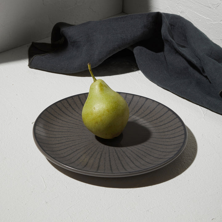 Ρηχό πιάτο Nordic της Estia σε τραπέζι με λευκή επιφάνεια και μαύρο λινό ύφασμα, διακοσμημένο με ένα πράσινο αχλάδι στο κέντρο.