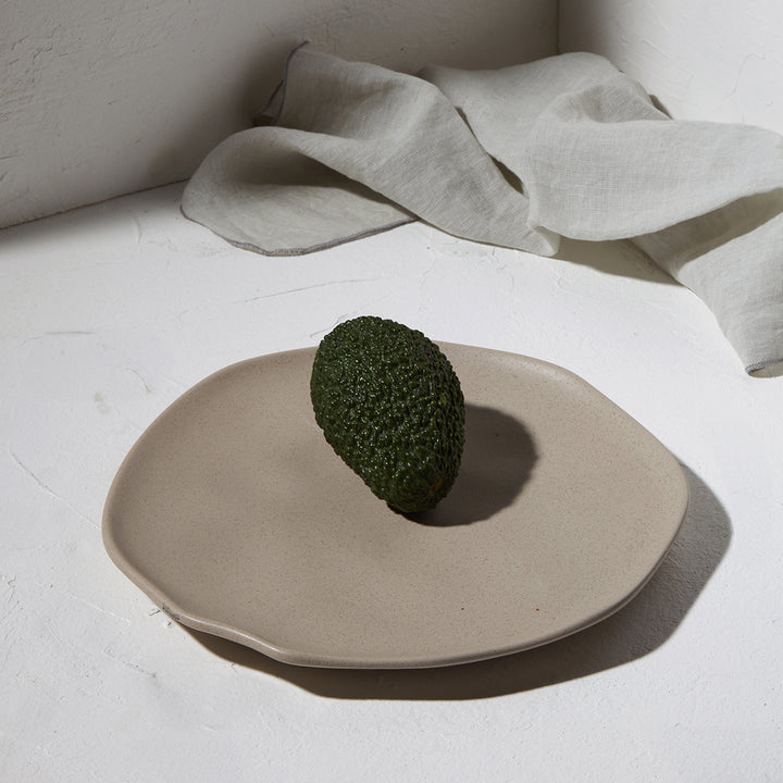 Πιάτο ρηχό Mare stoneware διάστασης 22cm της εταιρείας Estia, σε γκρι απόχρωση με ματ υφή, τοποθετημένο σε λευκό φόντο με υφασμάτινο διακοσμητικό και ένα αβοκάντο στο κέντρο.