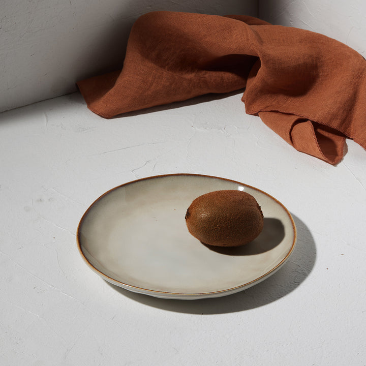 Ρηχό πιάτο Vignette της Estia τοποθετημένο σε λευκή επιφάνεια με αποξηραμένες φέτες πορτοκαλιού και ύφασμα στο φόντο.