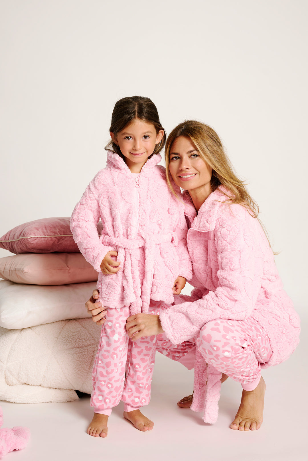 Μαμά και κόρη χαμογελούν φορώντας ροζ fleece ρόμπα που δένει με ζώνη και ανάγλυφα σχέδια καρδιές, συνδυασμένες με ροζ παντελόνι με μοτίβο leopar, σε ουδέτερο φόντο με μαξιλάρες λευκές και ροζ στο πλάι.