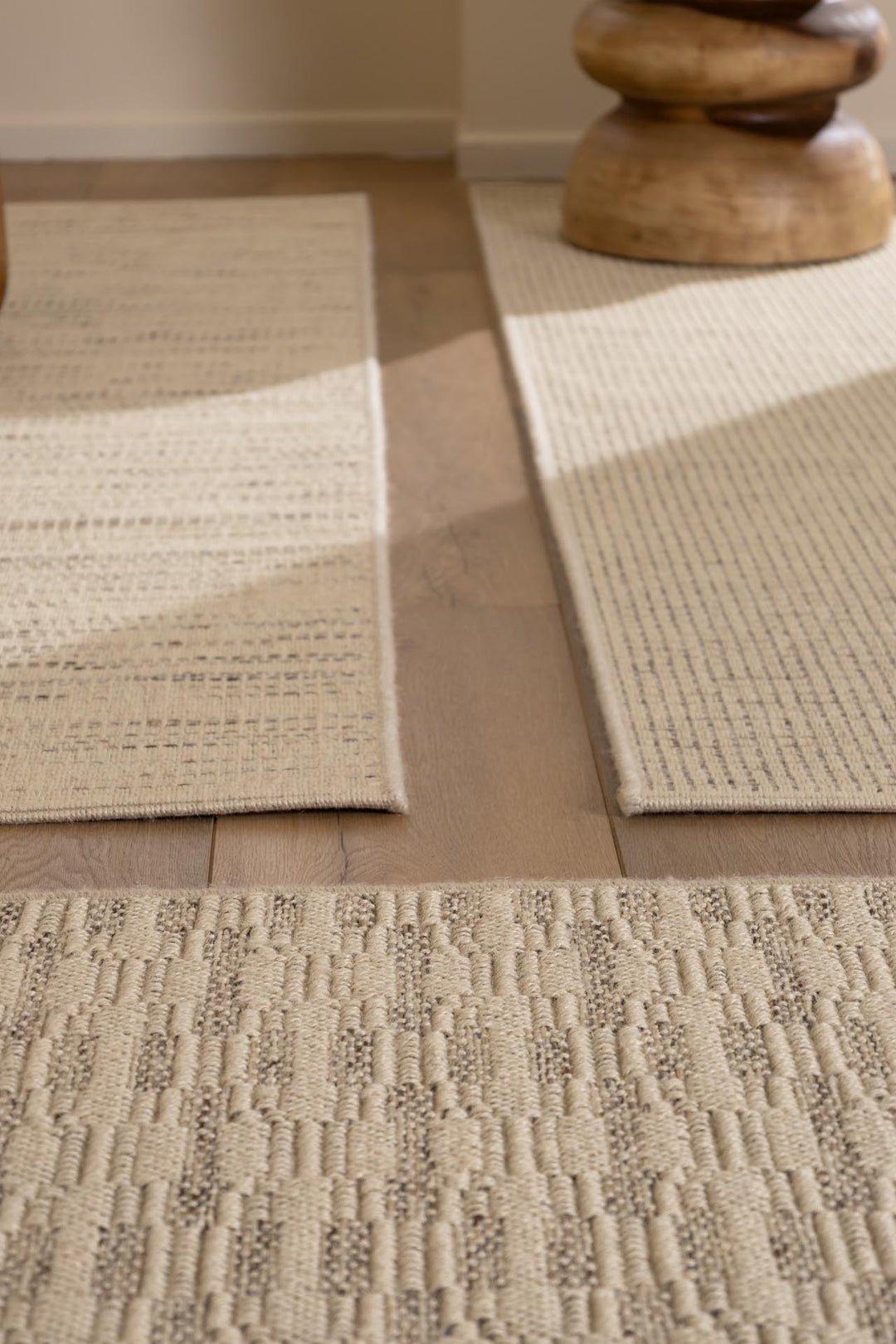 Σύνθεση με τρία μοντέρνα χαλιά σε απαλές μπεζ αποχρώσεις τοποθετημένα σε ξύλινο πάτωμα, με φυσικό φως και διακριτική σκίαση. Ιδανική πρόταση για φωτεινή, γήινη διακόσμηση σαλονιού ή καθιστικού.