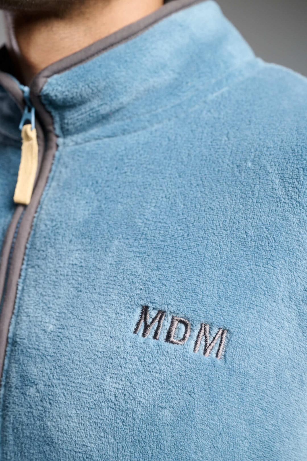 Λεπτομέρεια γαλάζιας fleece ζακέτας με όρθιο γιακά, γκρι φερμουάρ και κεντημένο λογότυπο MDM.