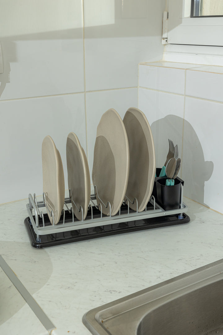 Ο στεγνωτήρας πιάτων τοποθετημένος σε μαρμάρινο πάγκο κουζίνας δίπλα στον νεροχύτη, περιλαμβάνοντας τέσσερα κεραμικά πιάτα και μαχαιροπίρουνα με γαλάζιες λαβές.