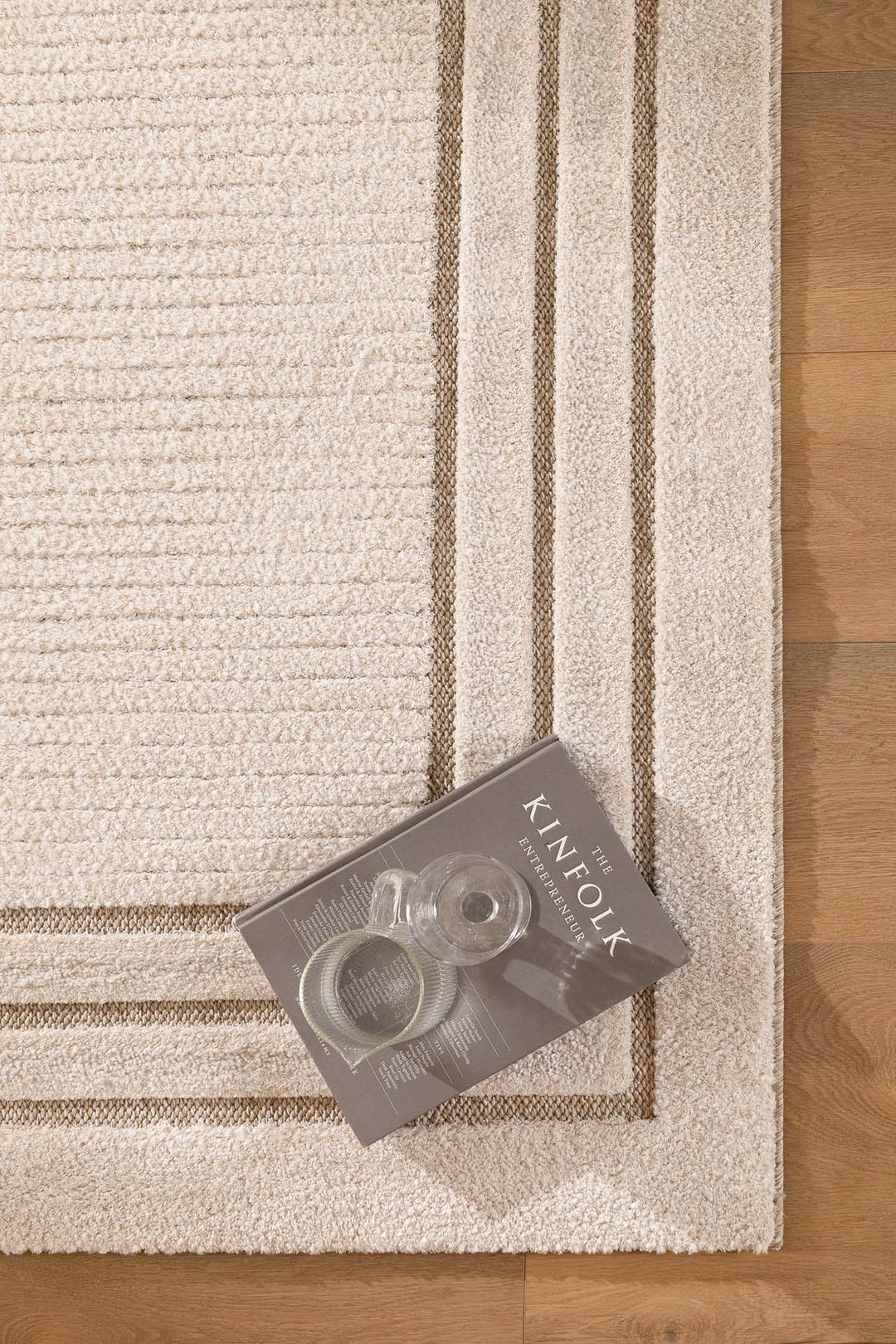 Μπεζ χαλί με γεωμετρικό πλαίσιο και υφή ριγέ, φωτογραφημένο δίπλα σε βιβλίο και ποτήρι πάνω σε ξύλινο πάτωμα.