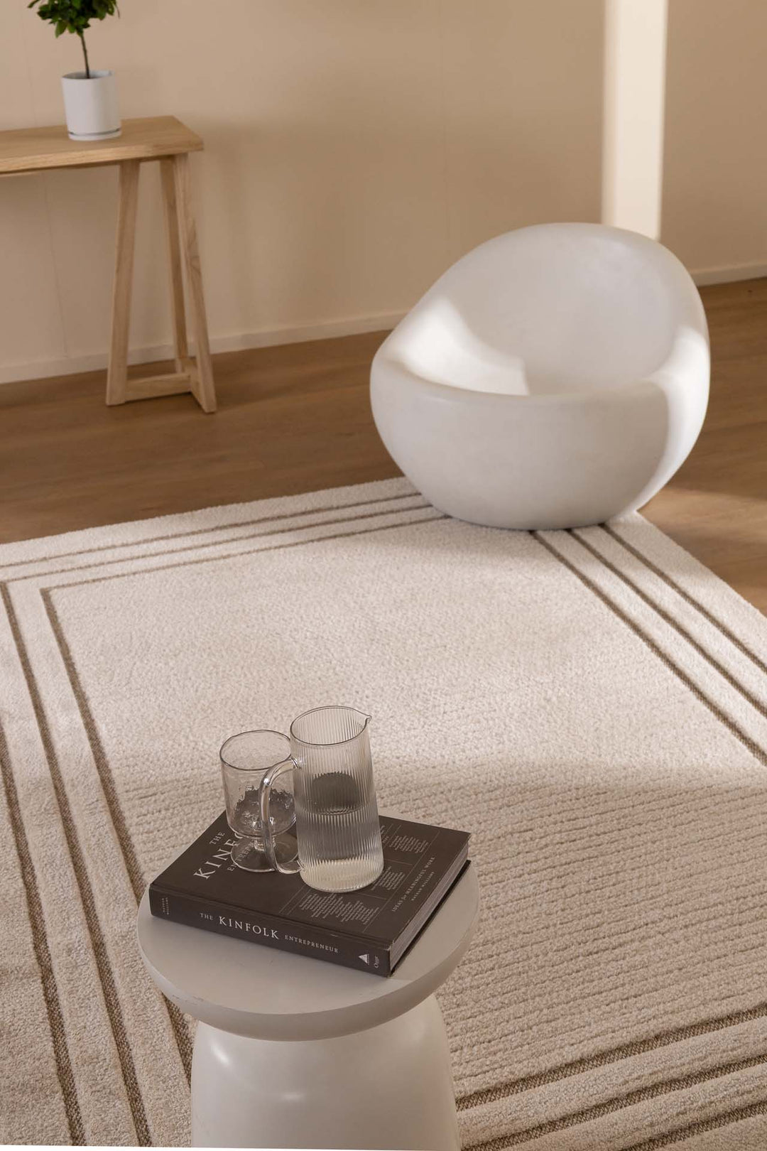 Minimal σαλόνι με μπεζ χαλί σε γήινες αποχρώσεις, λευκή πολυθρόνα και ξύλινο τραπεζάκι με διακοσμητικά.