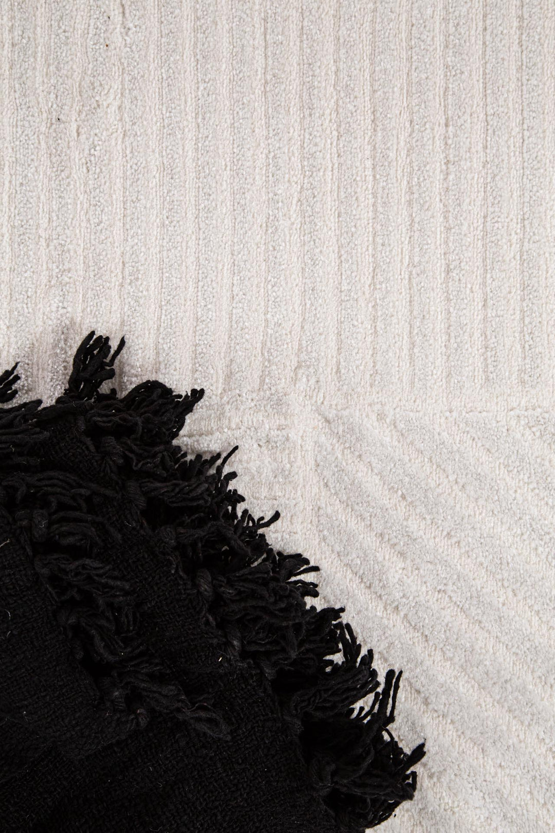 Λεπτομέρεια χαλιού Organic Royal Carpet σε cream χρώμα με ανάγλυφο γεωμετρικό σχέδιο, διακοσμημένο με μαύρη φουντωτή ριγέ κουβέρτα.