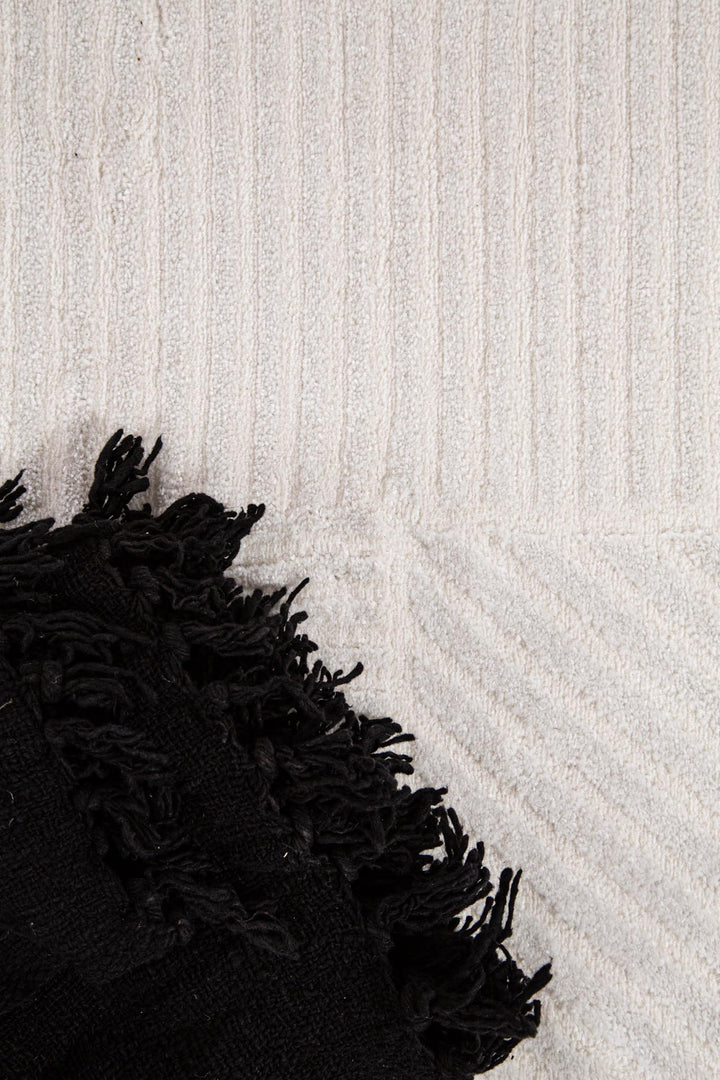 Λεπτομέρεια χαλιού Organic Royal Carpet σε cream χρώμα με ανάγλυφο γεωμετρικό σχέδιο, διακοσμημένο με μαύρη φουντωτή ριγέ κουβέρτα.