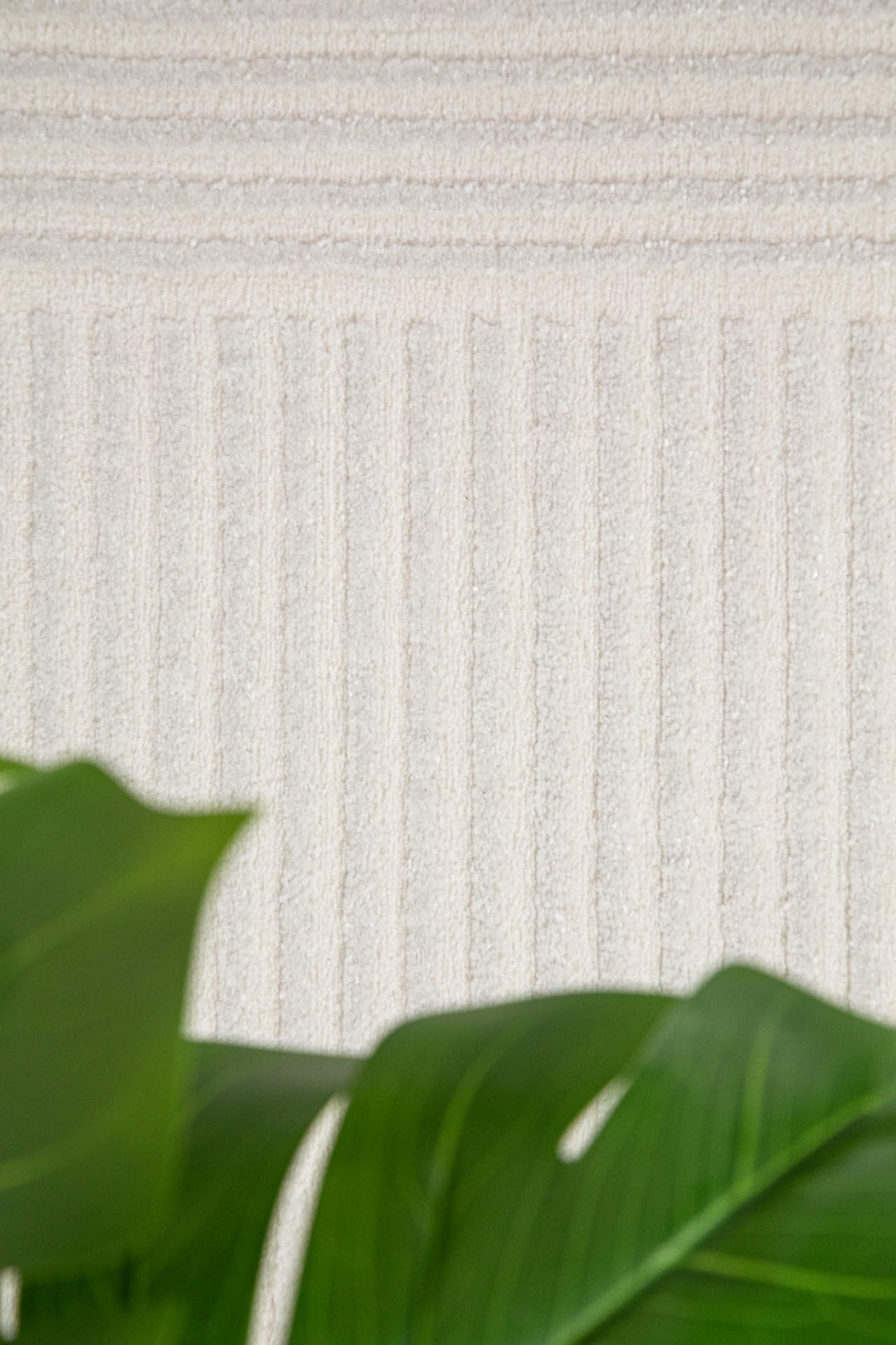 Κοντινό σε χαλί Organic της Royal Carpet σε cream απόχρωση με γεωμετρικό σχέδιο από κάθετες και οριζόντιες γραμμές, με πράσινο φύλλο στο φόντο.