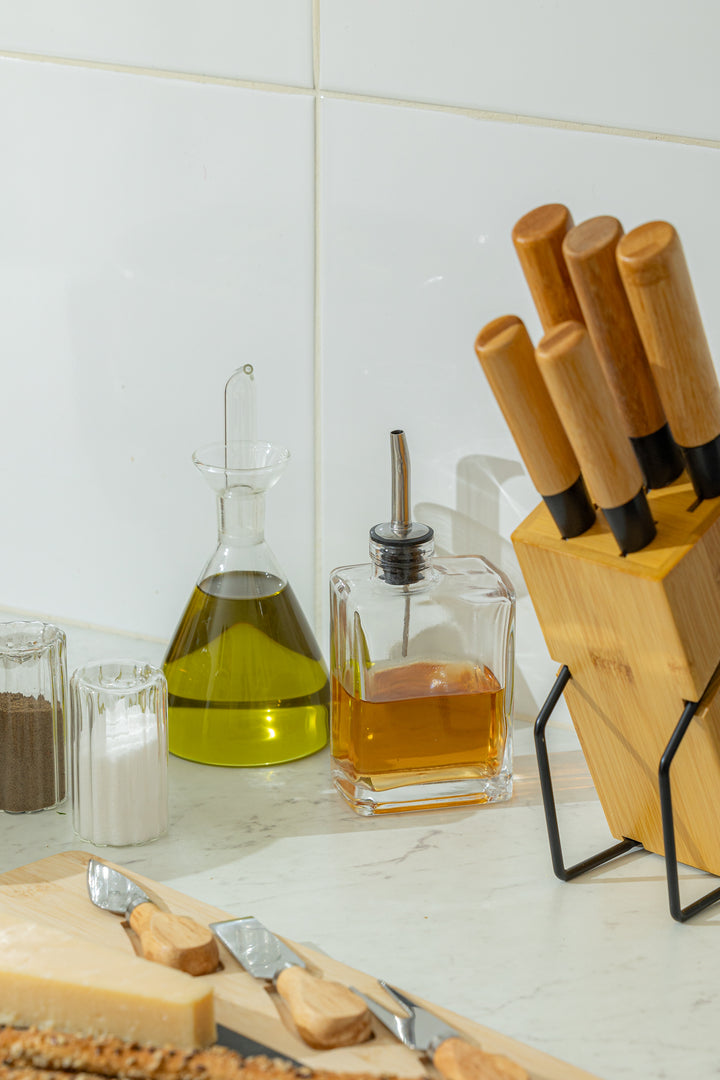 υάλινα δοχεία λαδιού και ξυδιού σε πάγκο κουζίνας, δίπλα σε μαχαίρια, ξύλινα αξεσουάρ και τυριά.