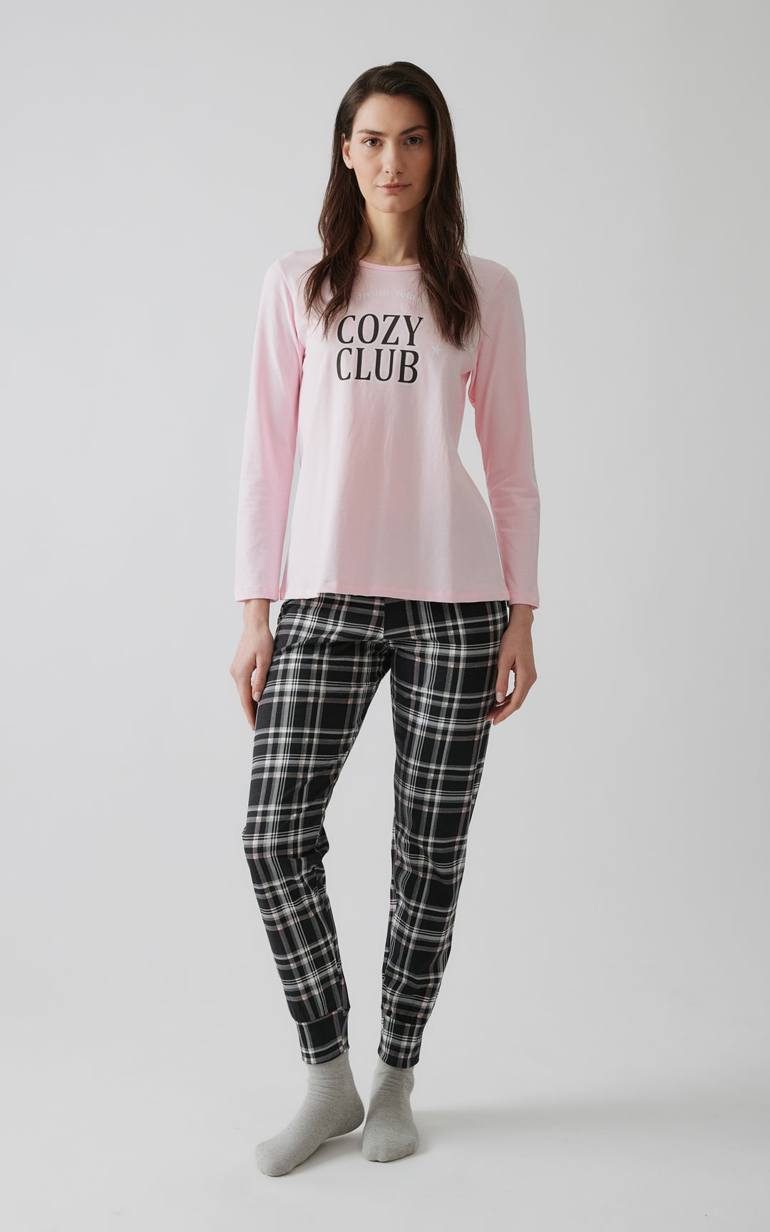 Γυναίκα ποζάρει σε στούντιο φορώντας ροζ μακρυμάνικη πιτζάμα με τύπωμα "COZY CLUB" και καρό παντελόνι σε μαύρο, λευκό και γκρι χρώμα, σε καθαρό φόντο με φυσικό φωτισμό.