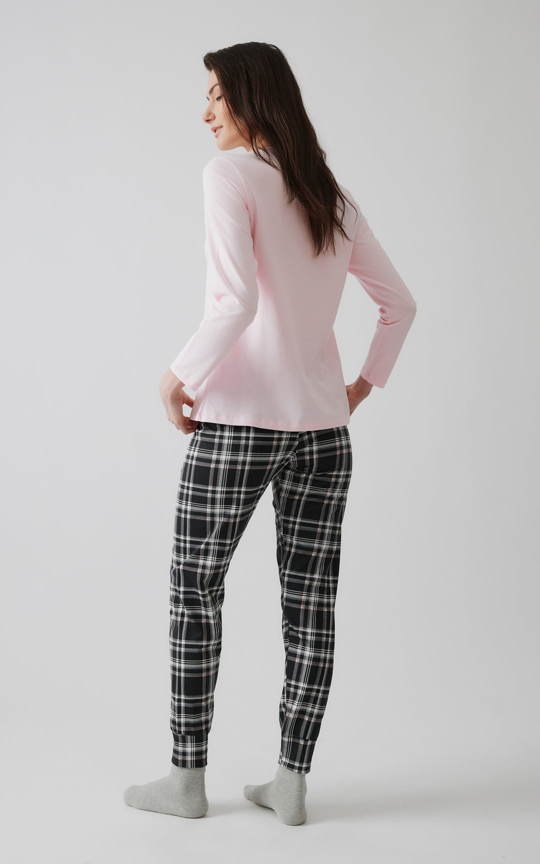Πλάγια όψη γυναίκας με ροζ μπλούζα και μαύρο-λευκό καρό παντελόνι πιτζάμας σε minimal στούντιο, που αναδεικνύει την απλότητα και τη χαλαρή γραμμή της πιτζάμας.