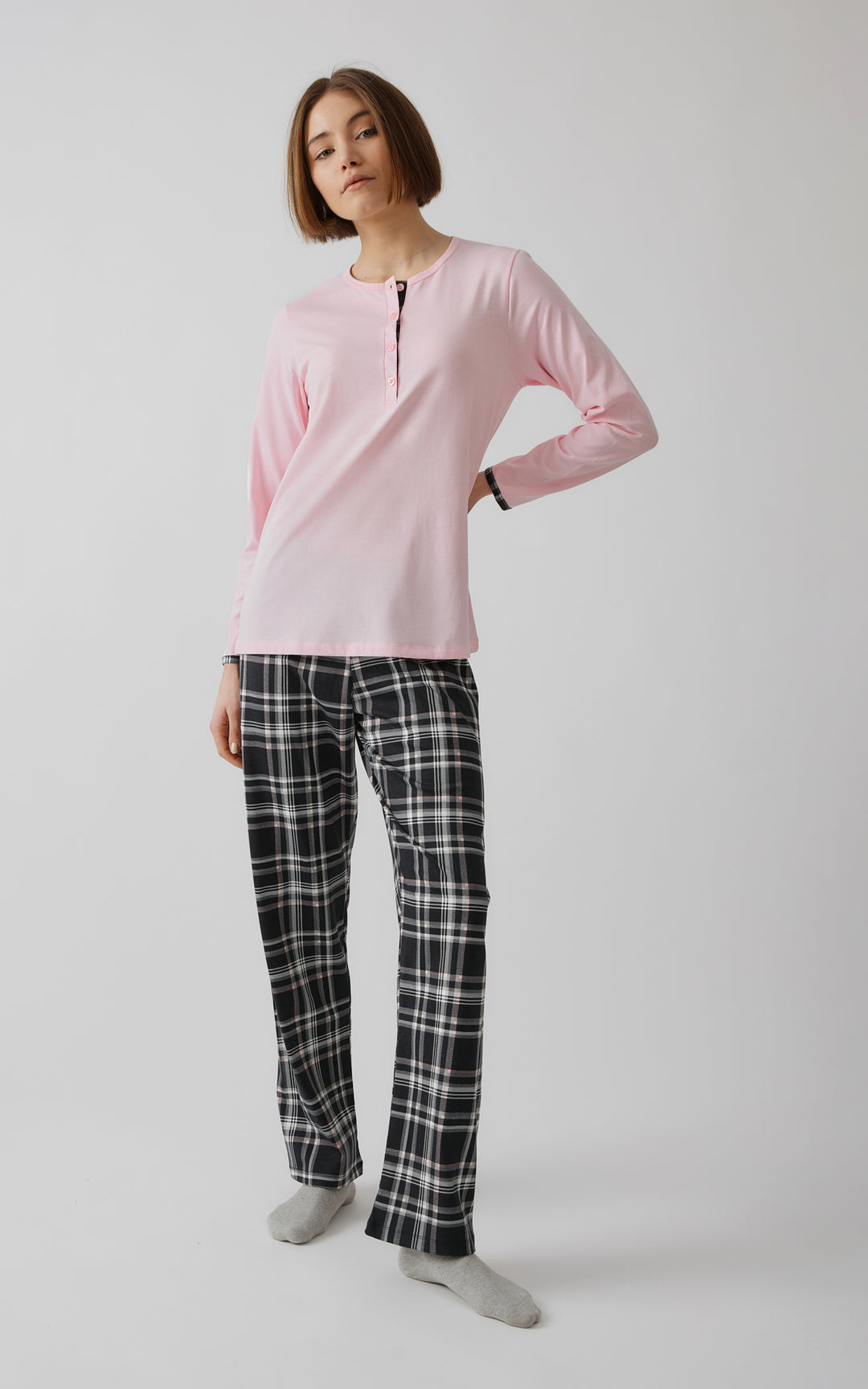 Γυναικείο μοντέλο μπροστά από γκρι φόντο ποζάρει με το ένα χέρι στη μέση και φορώντας ροζ πιτζάμα με κουμπάκια και καρό παντελόνι.