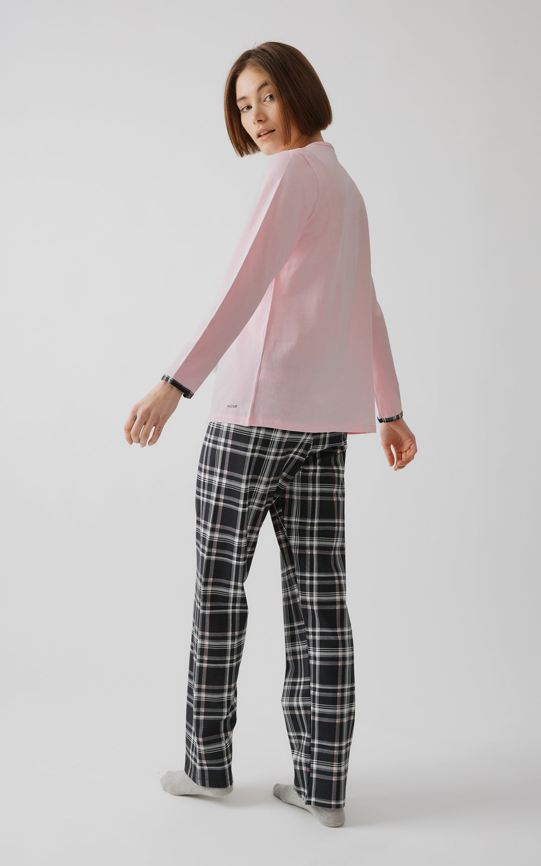 Γυναικεία πιτζάμα σε απαλό ροζ χρώμα με μαύρο καρό παντελόνι, σε στούντιο με λευκό φόντο. Το μοντέλο γυρίζει ελαφρώς προς τα πίσω, αναδεικνύοντας την πλάτη του σετ και τις λεπτομέρειες στους καρπούς.