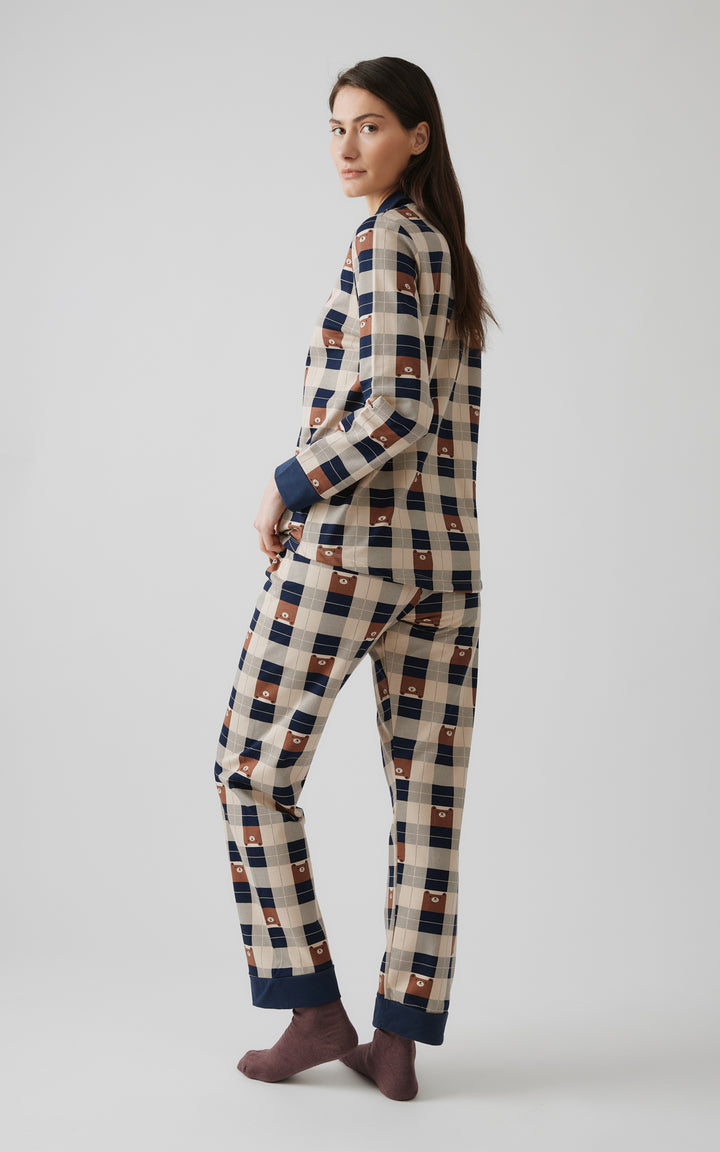 Γυναικείο μοντέλο γυρνά ελαφρώς προς τα πίσω, φορώντας καρό πιτζάμα σε γήινες και μπλε αποχρώσεις με διακριτικά σχέδια από αρκουδάκια και καφέ κάλτσες. 