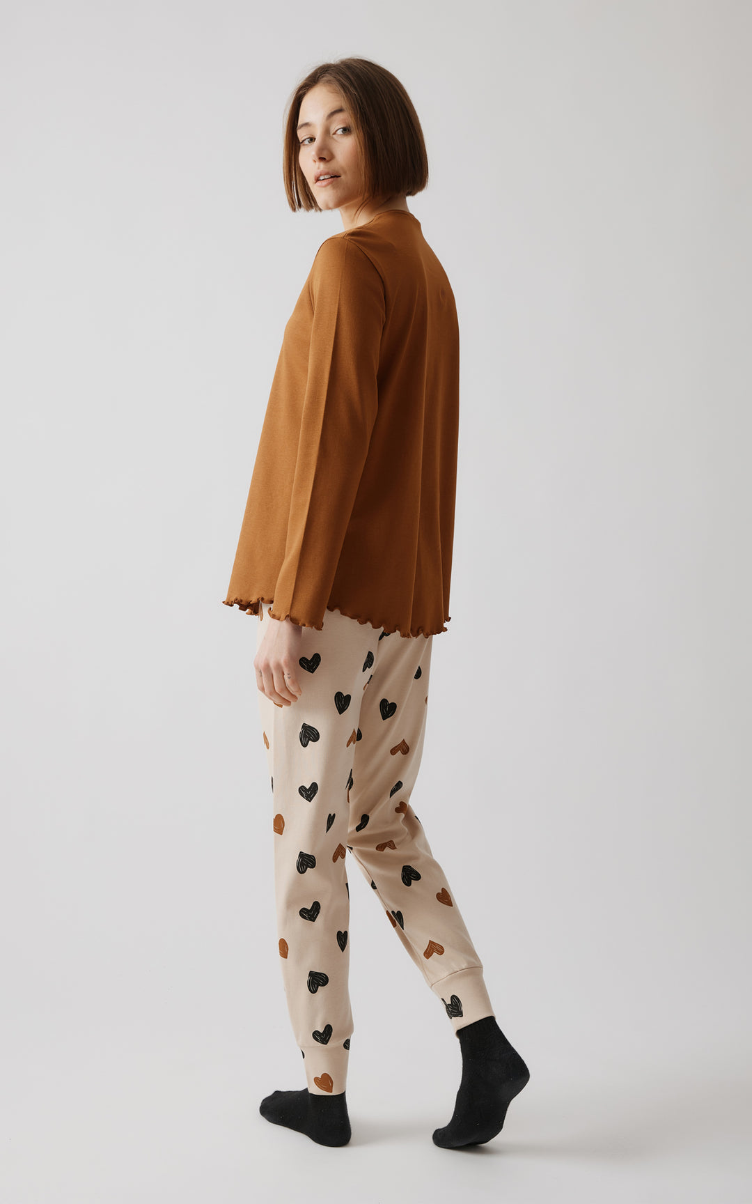 Πίσω όψη του γυναικείου μοντέλου με κοντά μαλλιά που φοράει χειμερινή καφέ πιτζάμα και παντελόνι με λάστιχο και print από καρδιές, σε γκρι φόντο.