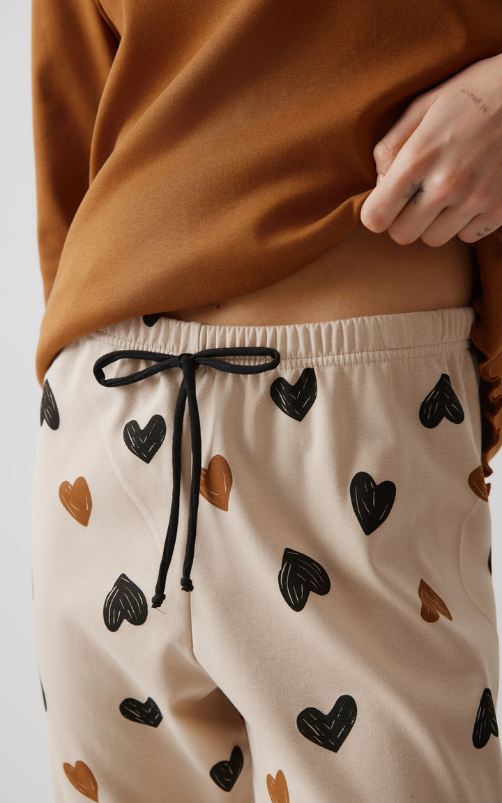 Κοντινό πλάνο που δείχνει το μπεζ παντελόνι πιτζάμας με σχέδια καρδιές και μαύρο κορδόνι στη μέση, ενώ το μοντέλο κρατά ελαφρώς τη μπλούζα.