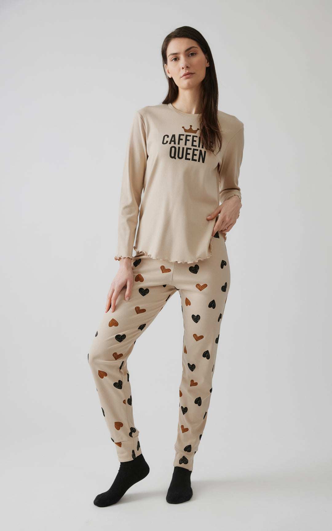 Γυναικείο μοντέλο ποζάρει με χαλαρό βλέμμα φορώντας μπεζ πιτζάμα με στάμπα "CAFFEINE QUEEN" και παντελόνι με καφέ και μαύρες καρδιές, σε γκρι φόντο.
