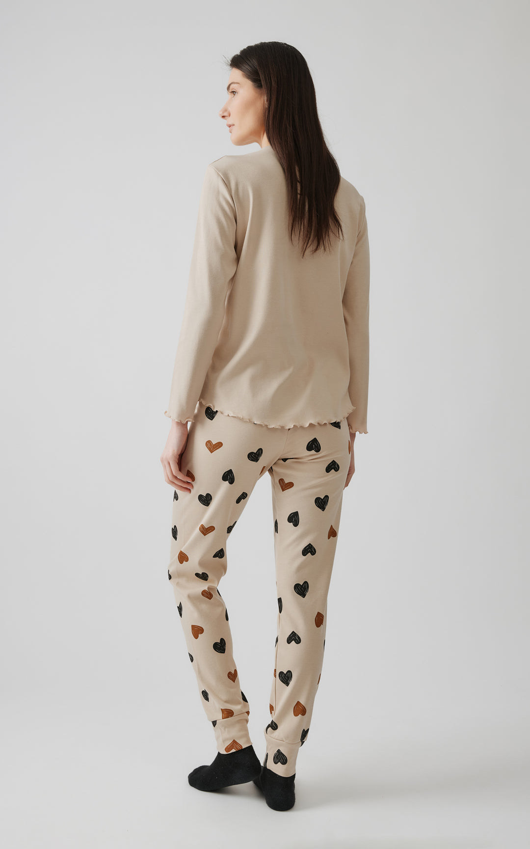 Γυναικείο μοντέλο ποζάρει γυρισμένο πλάτη προς την κάμερα, αναδεικνύοντας τη συνολική όψη της μπεζ πιτζάμας με σχέδια καρδιές, μπροστά σε γκρι φόντο.