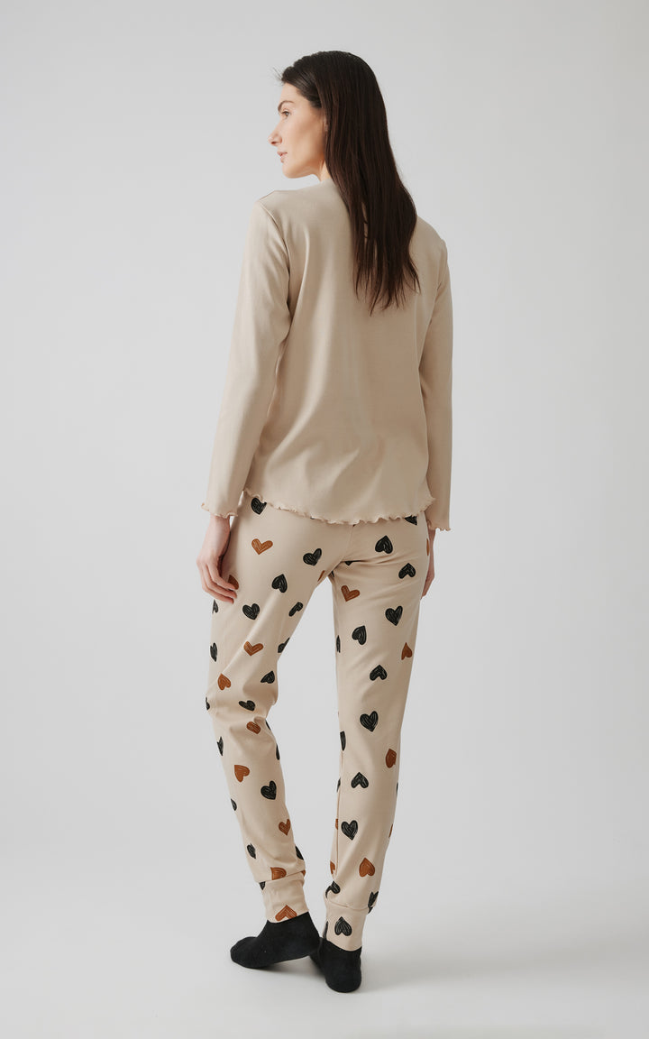 Γυναικείο μοντέλο ποζάρει γυρισμένο πλάτη προς την κάμερα, αναδεικνύοντας τη συνολική όψη της μπεζ πιτζάμας με σχέδια καρδιές, μπροστά σε γκρι φόντο.