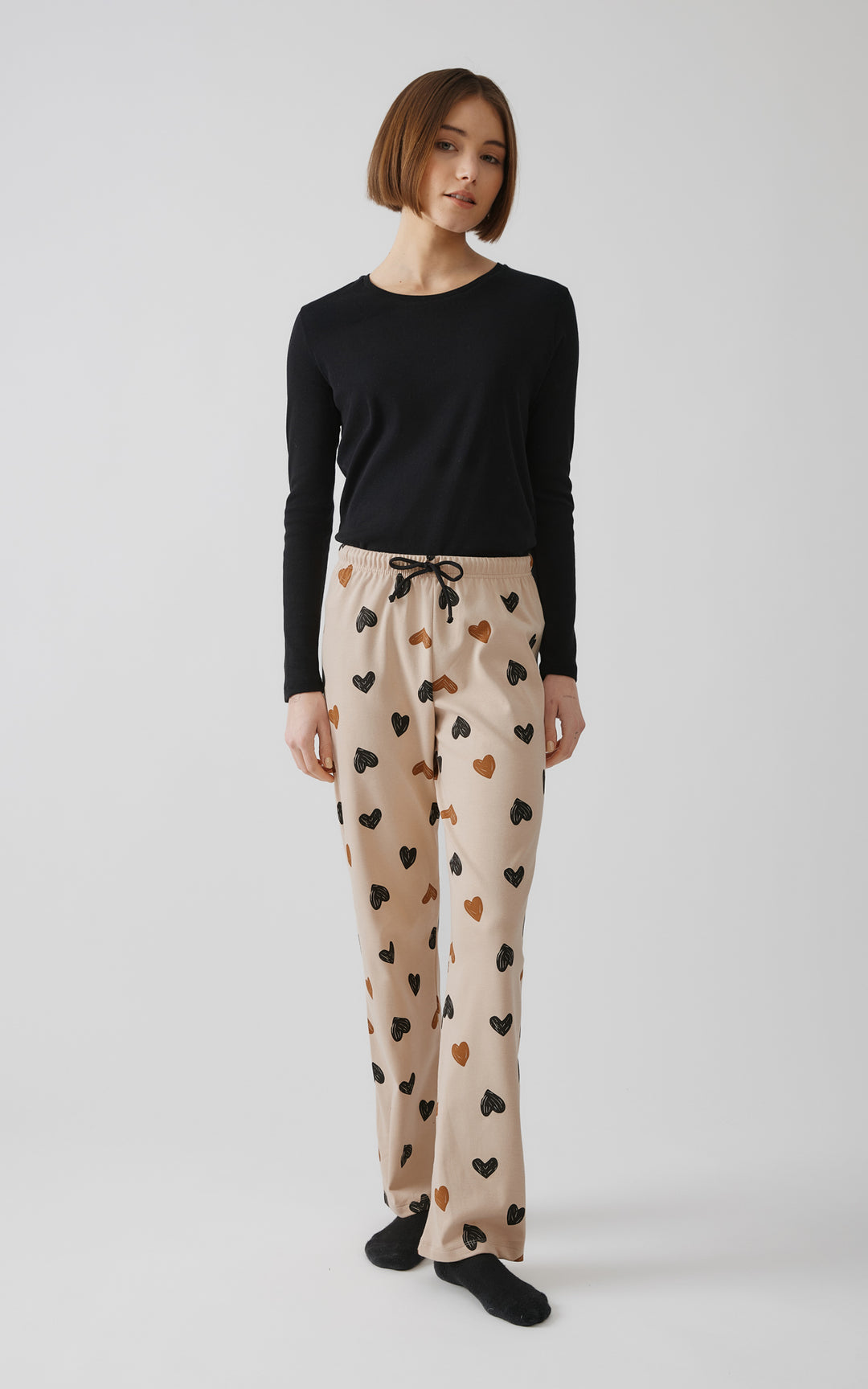 Γυναικεία πιτζάμα με μαύρη μπλούζα μακρυμάνικη και μπεζ παντελόνι με μοτίβο καρδιές, ολόσωμη λήψη σε λευκό φόντο.